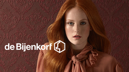 Still uit De Bijenkorf commercial met logo en vrouw met rood haar op achtergrond.